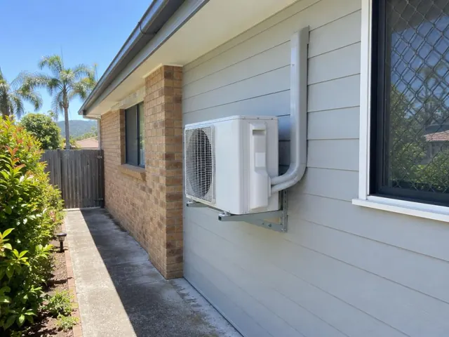 Outdoor air conditioning unit mounted on a wall beside a brick home in Helensvale